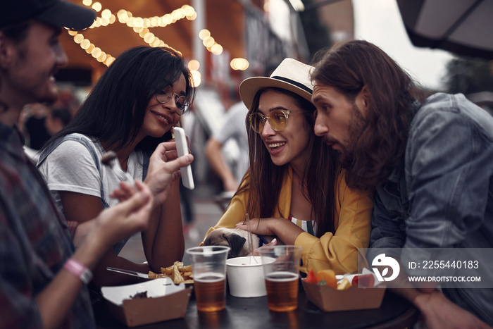 Check this out. Portrait of young people standing at the table and watching funny video on cellphone. Lovely girl in hat holding pug dog