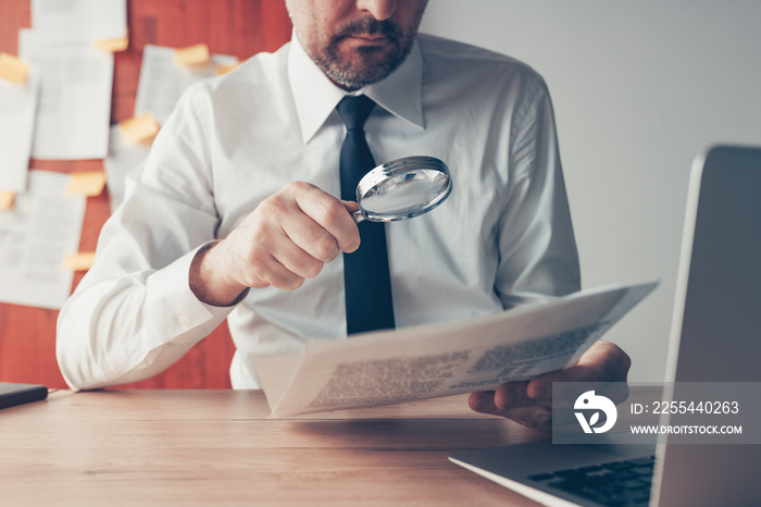 Businessman reading document contract papers with magnifying glass at office desk