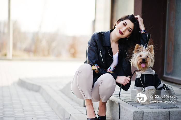 Brunette gypsy girl with yorkshire terrier dog posed against large windows house. Model wear on leather jacket and t-shirt with ornament, pants and shoes with high heels.