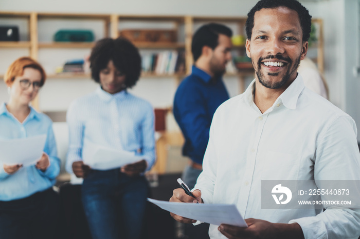 Teamwork concept in modern office.Young african businessman wearing white shirt holding papers at hands and standing front of the coworkers team.Horizontal,blurred background.