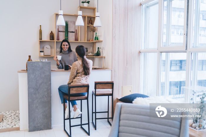 Cheerful girlfriends communicating while sitting at counter with cups of hot drink at home