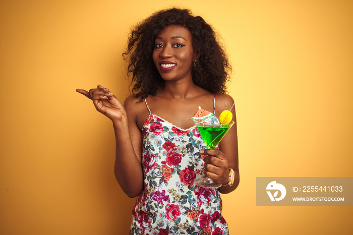 Young african american woman drinking cocktail standing over isolated yellow background very happy pointing with hand and finger to the side