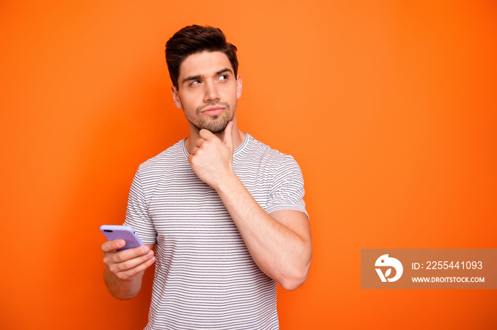 Photo of minded serious millennial guy hold telephone deep think new instagram blog post text arm on chin look side wear striped t-shirt isolated bright orange color background