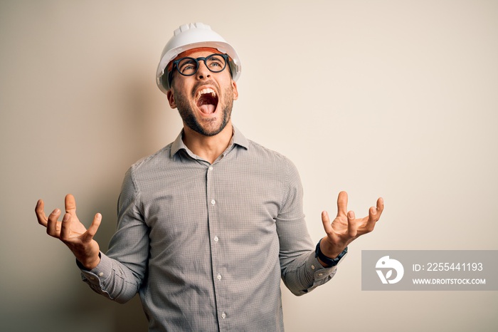 Young architect man wearing builder safety helmet over isolated background crazy and mad shouting and yelling with aggressive expression and arms raised. Frustration concept.