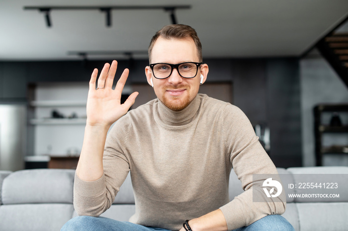 Young adult European man in elegant black glasses, looking at the camera, smiling and waving the hand, say hello, greeting by video, speaking on webcam, positive person, with white earphones, stylish