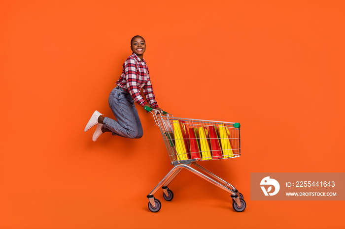 Full body photo of carefree excited girl jumping push trolley have good mood isolated on orange color background