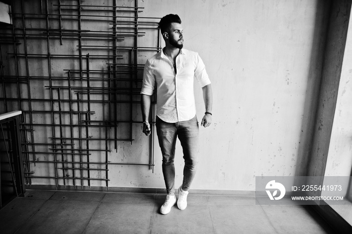 Stylish tall arabian man model in white shirt, jeans posed against steel wall indoor. Beard attractive arab guy.