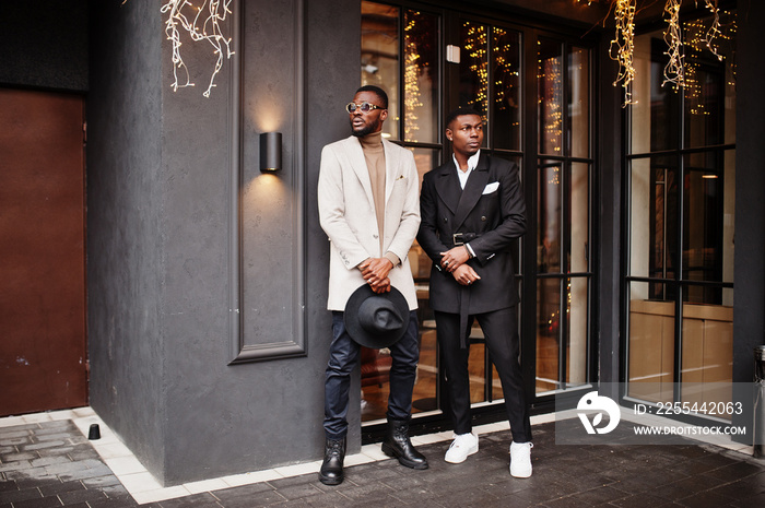 Two fashion black men pose against house with garlands. Fashionable portrait of african american male models. Wear suit, coat and hat.