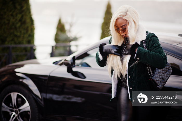 Chic african american blond woman in black leather clothes posed against race modern car.