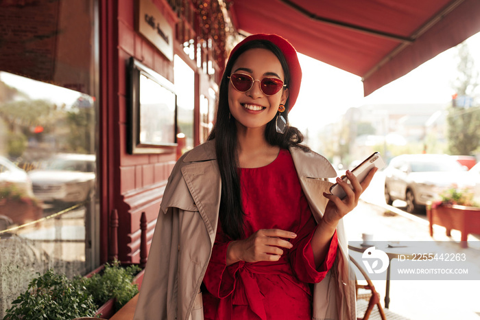 Tanned young brunette woman smiles sincerely in street cafe. Attractive Asian lady in red beret, sunglasses, stylish dress and trendy trench coat holds phone.