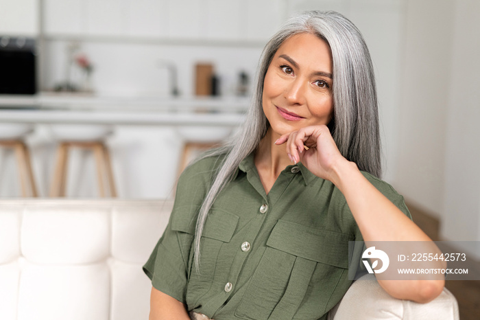 Close-up portrait of charming middle-aged woman with long silver hair sitting in relaxed athmosphere, mature 50s asian female resting at home, looking at camera and smiling with calm smile