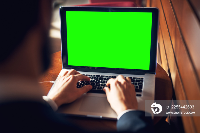 Close up rear view of young bearded businessman hands typing on a laptop keyboard with a blank green editable screen near the wooden wall.