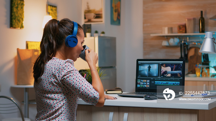 Woman video editor with headset working with footage and sound sitting in home kitchen. Woman videographer editing audio film montage on professional laptop sitting on desk in midnight
