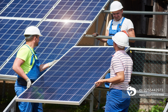 Team of three young technicians in protective helmets mounting heavy solar photo voltaic panel on metal platform. Exterior solar panel system installation, efficiency and durability concept.