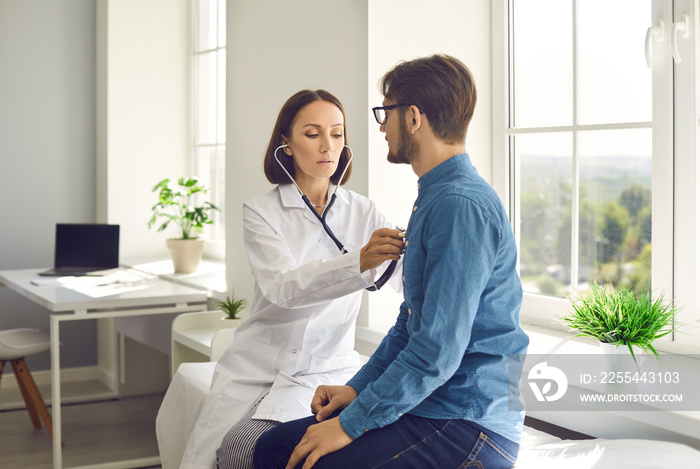 Young man visiting clinic to check heart or lungs health. Serious female doctor sitting on examination couch in modern exam room, holding stethoscope, listening to male patient’s breath or heartbeat