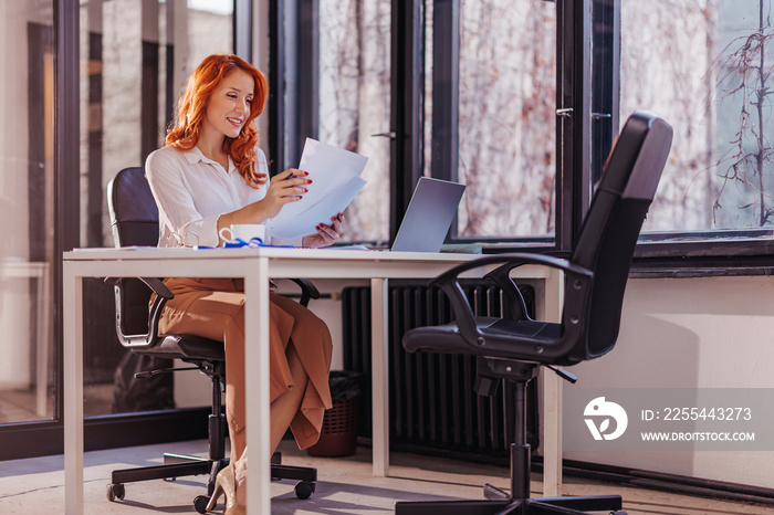 Happy smiling businesswoman in formal wear sitting at desk in modern office in front of laptop and doing paperwork