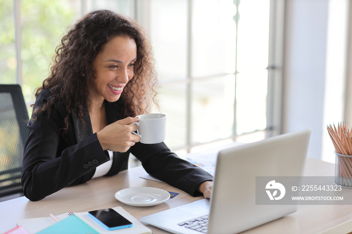 Attractive professional latin female employee worker sitting, using laptop computer while drinking coffee at home workplace. Businesswoman working on paperworks and communicate with client.