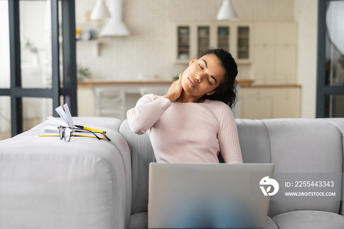 African American female freelancer or student with curly hair sitting on the couch in modern living room, feeling tired from constant work on laptop, removed her glasses, stretching neck with hand