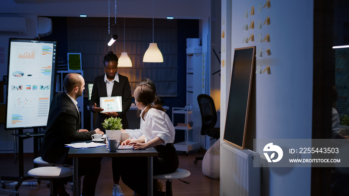 Overworked workaholic businesswoman with dark skin explaining management graphs using tablet overworking in company meeting office room late at night. Multi-ethnic teamwork solving company problem