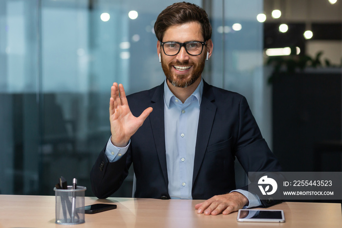 Video call on online meeting with colleagues, mature boss in business suit looking at web camera and smiling waving to employees, businessman in glasses working inside modern office building.