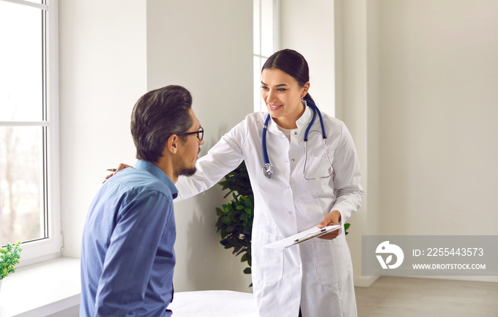 Professional female doctor approvingly, carefully puts her hand on the shoulder of young male patient. Handsome guy with glasses at doctor’s appointment in modern office of medical institution.