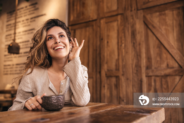 Joyful caucasian young woman sits at table in cafe and holds cup of coffee. Blonde girl drinks hot latte, looking away. Vacation concept