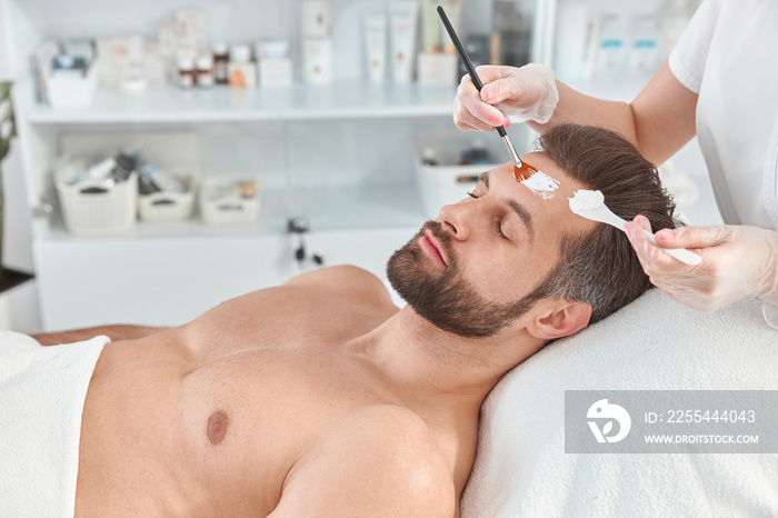 Bearded young man is relaxing while cosmetologist is spreading white clay on his face with brush at beauty salon