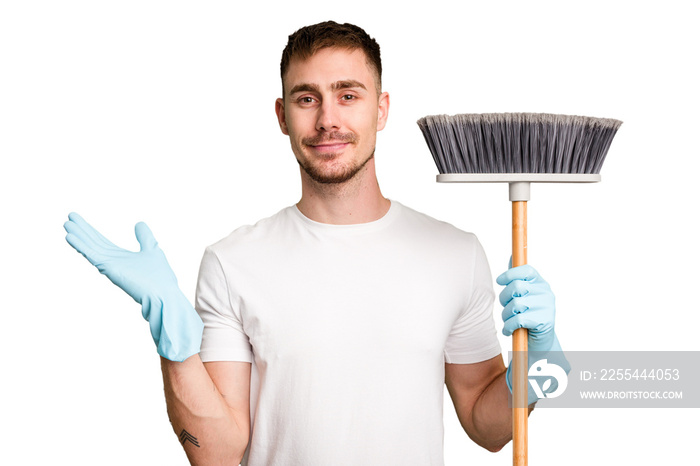 Young man holding a broom to clean his house cut out isolated showing a copy space on a palm and holding another hand on waist.