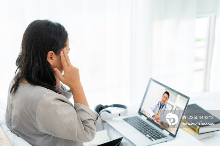 Asian woman in white bed speaking with doctor using tele health technology while sitting on a sofa and headache in bedroom at home while she sick during self quarantine for COVID-19.