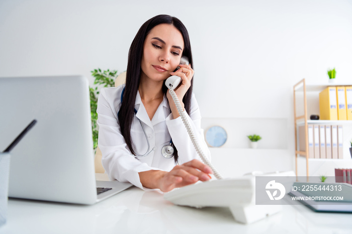 Photo of beautiful doctor serious lady modern technology notebook on table speaking telephone consulting clients wear white lab coat shirt sitting chair office clinic indoors