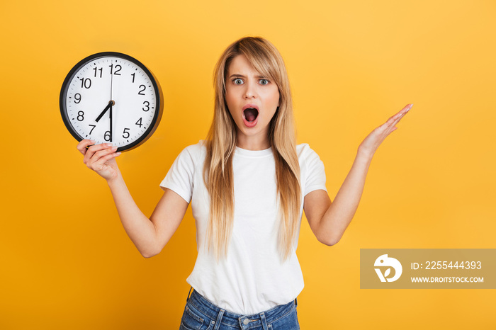 Shocked displeased young blonde woman posing isolated over yellow wall background dressed in white casual t-shirt holding clock.