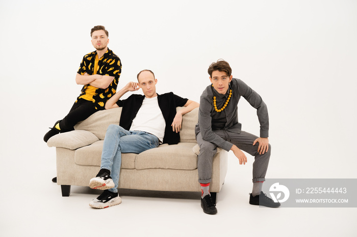 Friends seated on couch in studio. Three stylish young guys talking on the camera. Group of friends are sitting on a soft couch and communicates isolated on white background.