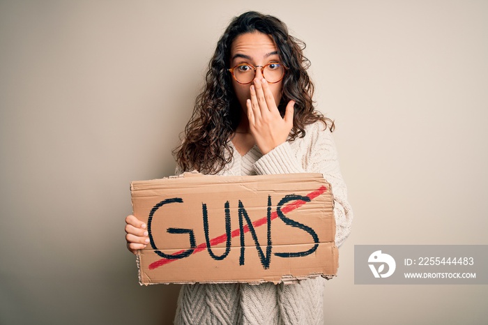 Young beautiful woman with curly hair holding banner with prohibited guns message cover mouth with hand shocked with shame for mistake, expression of fear, scared in silence, secret concept