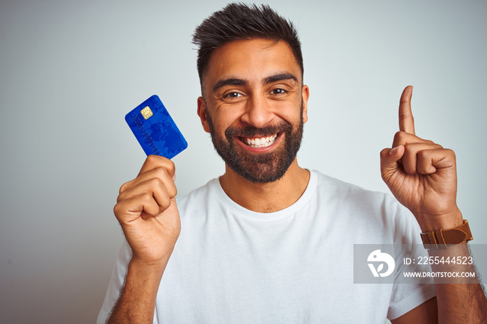 Young indian customer man holding credit card standing over isolated white background surprised with an idea or question pointing finger with happy face, number one