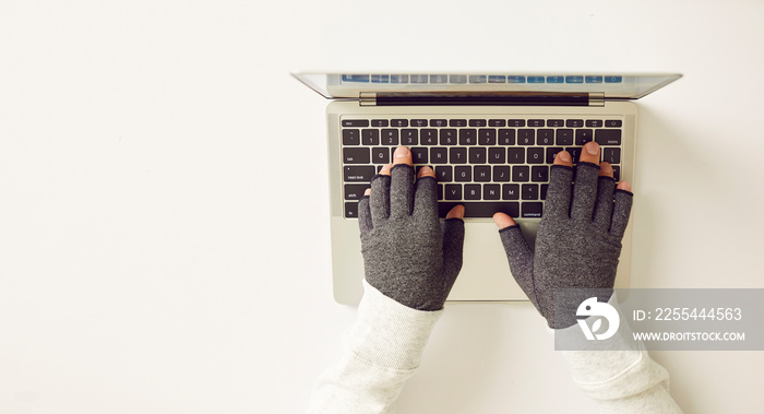 Man with rheumatoid arthritis wearing pair of warm comfortable open finger grey textile pain easing compression hand gloves working at white office desk on his notebook PC computer. Top view. Banner