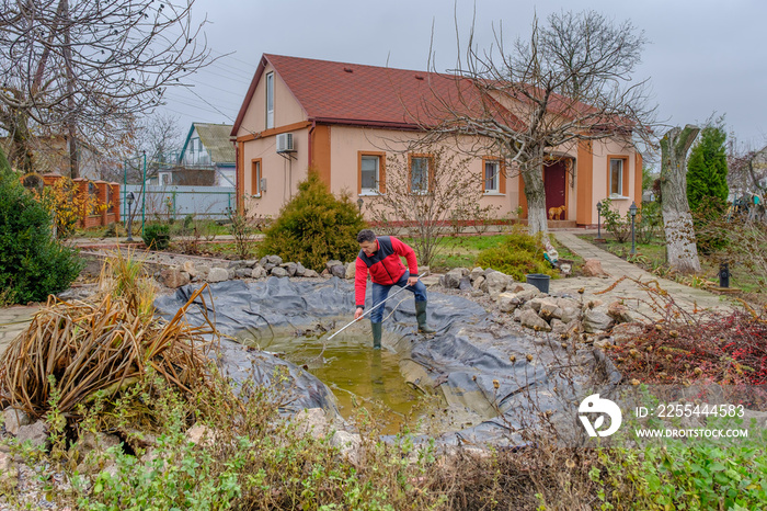 white man cleans a garden pond with a landing net from slime, water plants, falling leaves and catches fish for resettlement in an aquarium near his house. Autumn seasonal pond care before winter