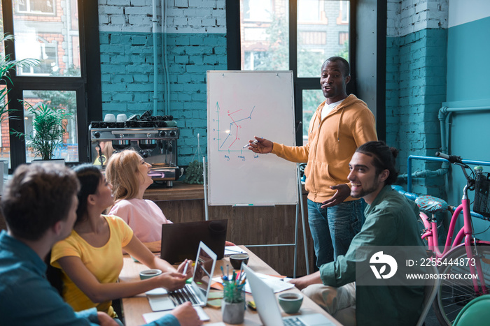 Portrait of outgoing male employee showing diagram on white board while standing near it. Cheerful colleagues looking at him. Occupation concept