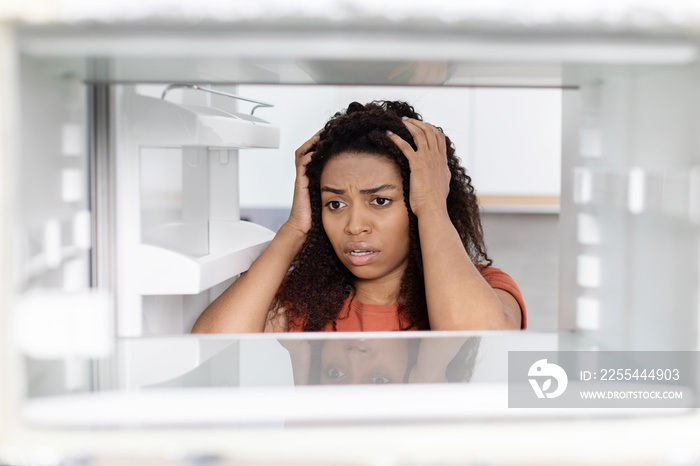 Sad shocked hungry millennial black lady hold her head, looking into empty refrigerator, suffering from starvation