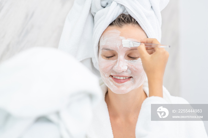 Young girl puts white cream on her mom’s face at home. Mom and child girl are in bathrobes and with towels on their heads