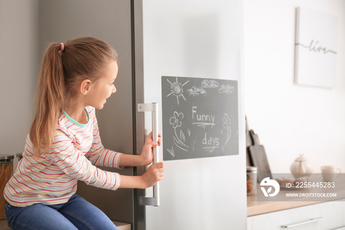 Little girl near chalkboard on refrigerator in kitchen