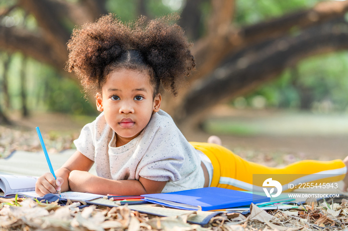 African American little girl lying drawing in the coloring book for kids and looking at camera in the park