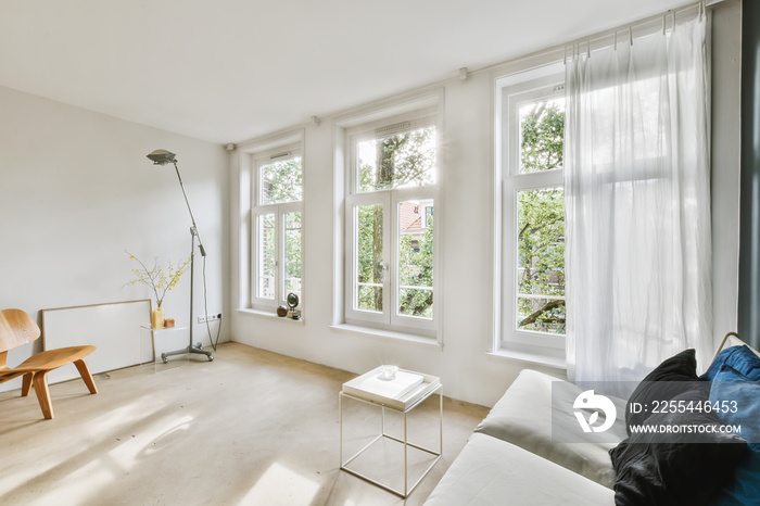 a living room with white walls and wood flooring, two large windows in the wall is covered with sheer curtains