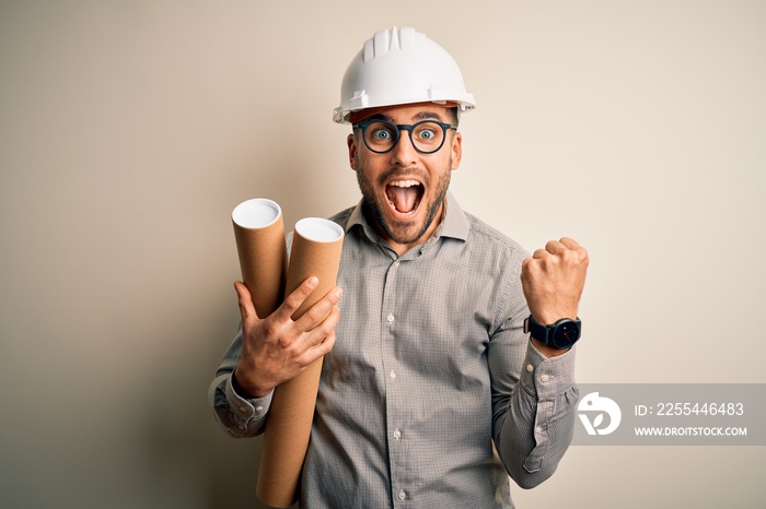 Young architect man wearing contractor helmet holding project paper plan over isolated background screaming proud and celebrating victory and success very excited, cheering emotion