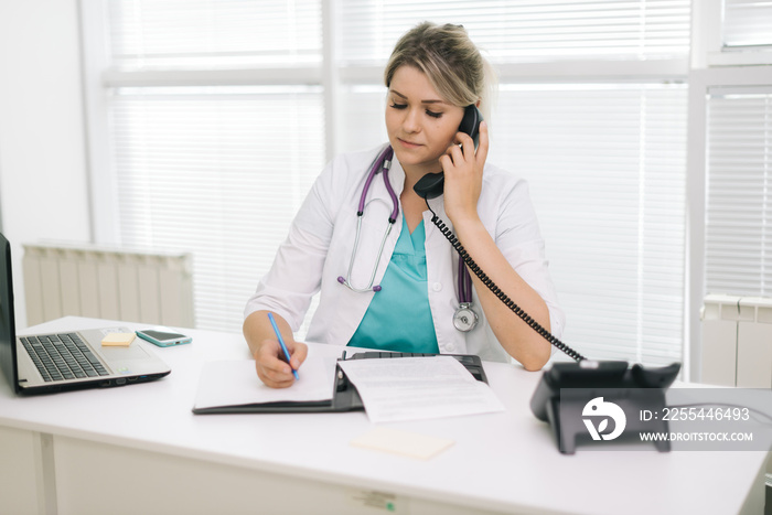 Portrait of young woman doctor in white uniform sitting at the table and writing a prescription in document while talking on the phone at office. Girl doc sitting against the background of a window