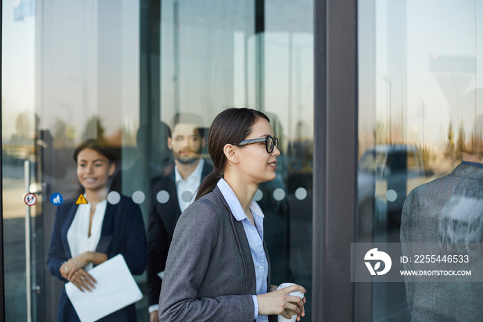 Smiling attractive young businesswoman in glasses holding coffee cup and walking through revolving door while coming in office building