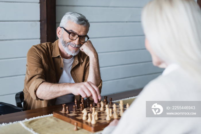 Smiling elderly man playing chess with wife on blurred foreground