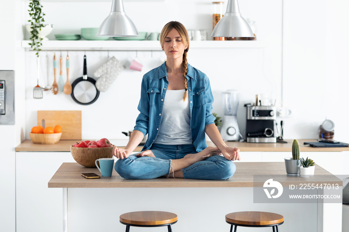 Pretty young woman in lotus position sitting on the table while relaxing in the kitchen at home.