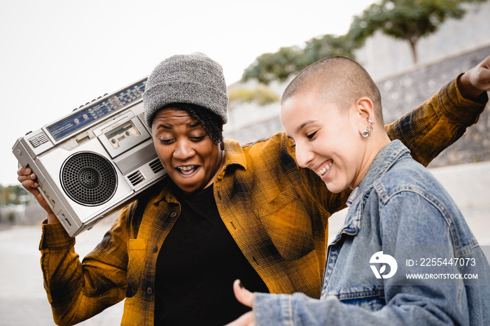 Happy multiracial people dancing to urban music outdoors with city on background - Women from diverse cultures celebrating together