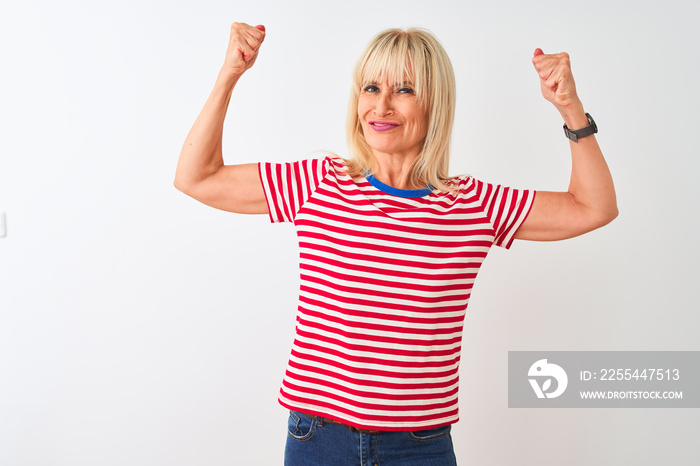 Middle age woman wearing casual striped t-shirt standing over isolated white background showing arms muscles smiling proud. Fitness concept.