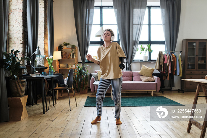 Young woman with prosthetic arm dancing while listening to music in wireless headphones in living room at home